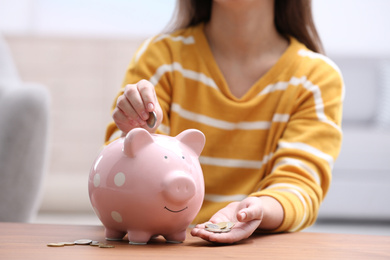 Woman putting coin into piggy bank at wooden table, closeup