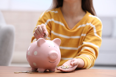 Photo of Woman putting coin into piggy bank at wooden table, closeup