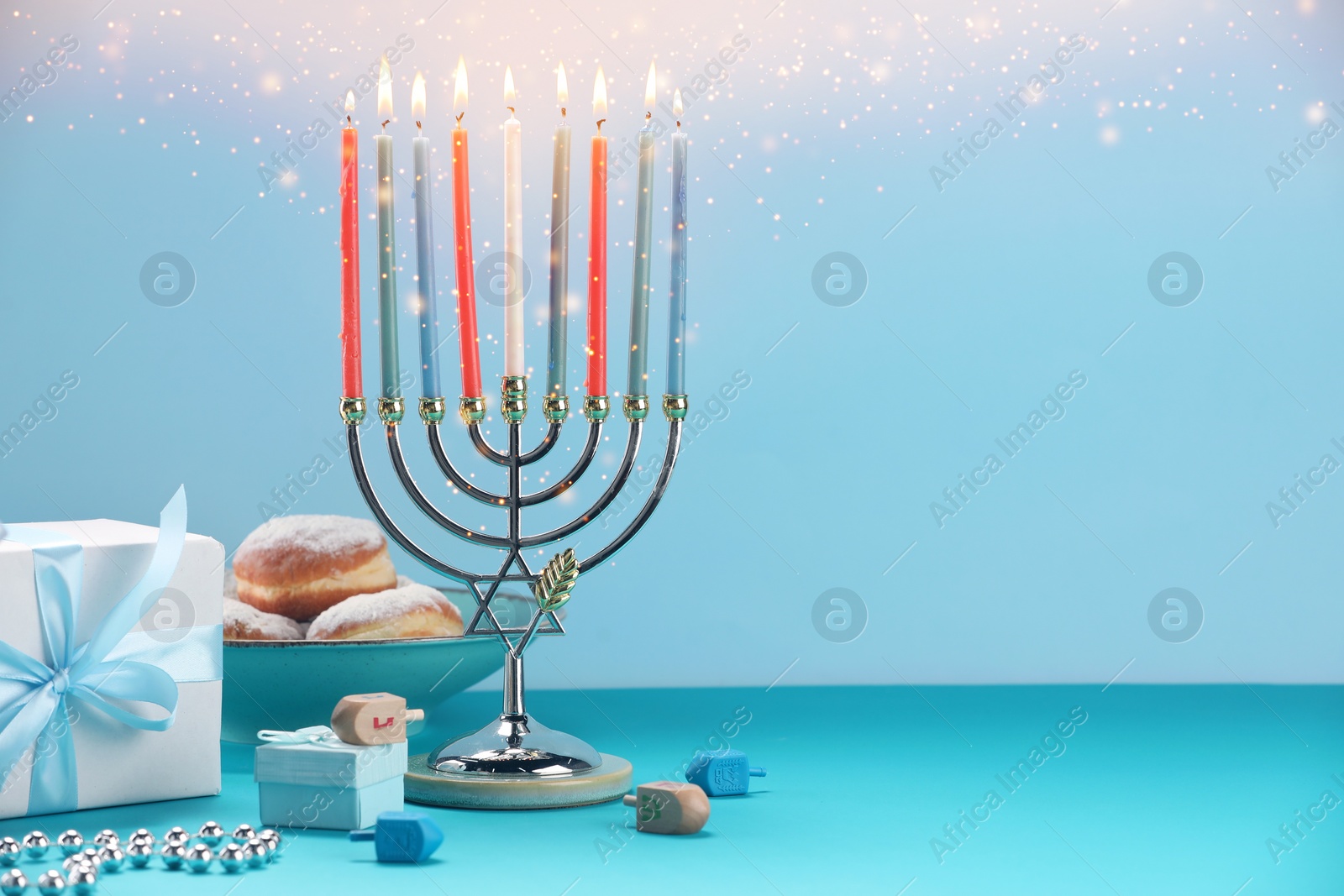 Image of Hanukkah celebration. Menorah with burning candles, dreidels, gift boxes and donuts on light blue table, space for text