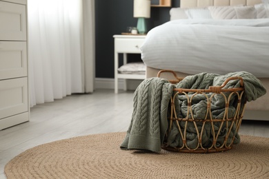 Photo of Knitted green plaid in wicker basket near bed indoors, space for text