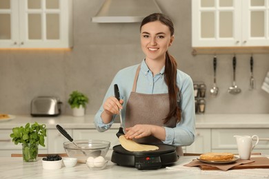 Photo of Happy woman cooking delicious crepe on electric maker at white marble table in kitchen