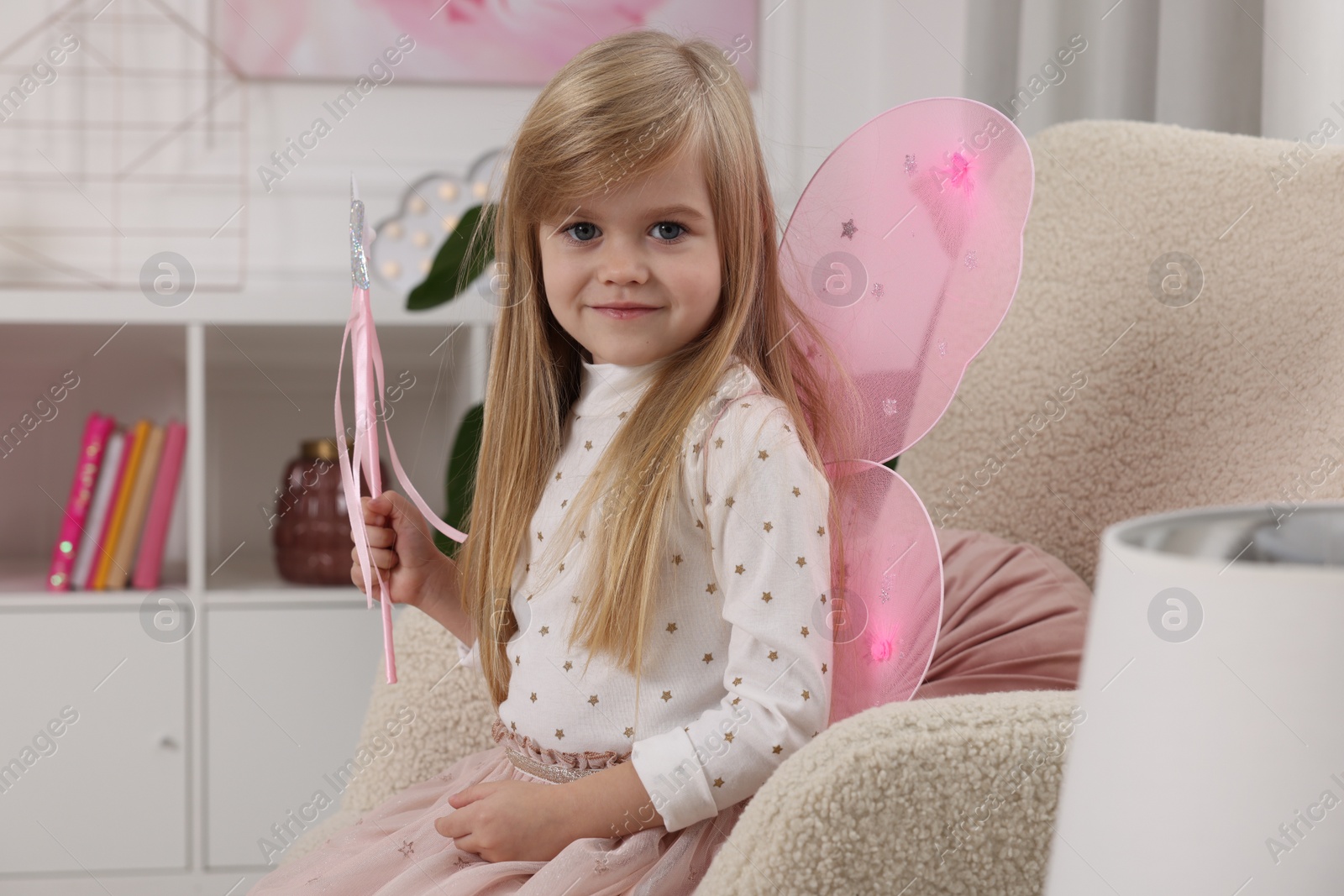 Photo of Cute little girl in fairy costume with pink wings and magic wand in armchair at home