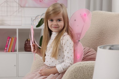 Photo of Cute little girl in fairy costume with pink wings and magic wand in armchair at home