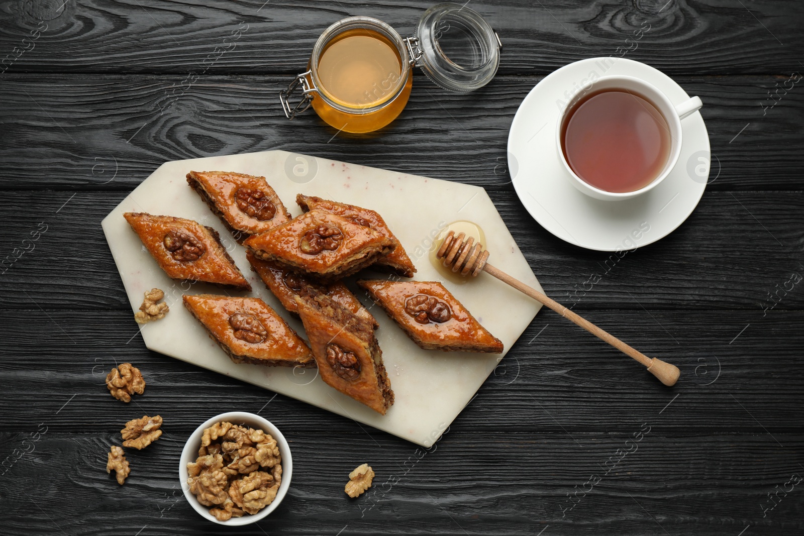 Photo of Delicious sweet baklava with walnuts, honey and hot tea on black wooden table, flat lay