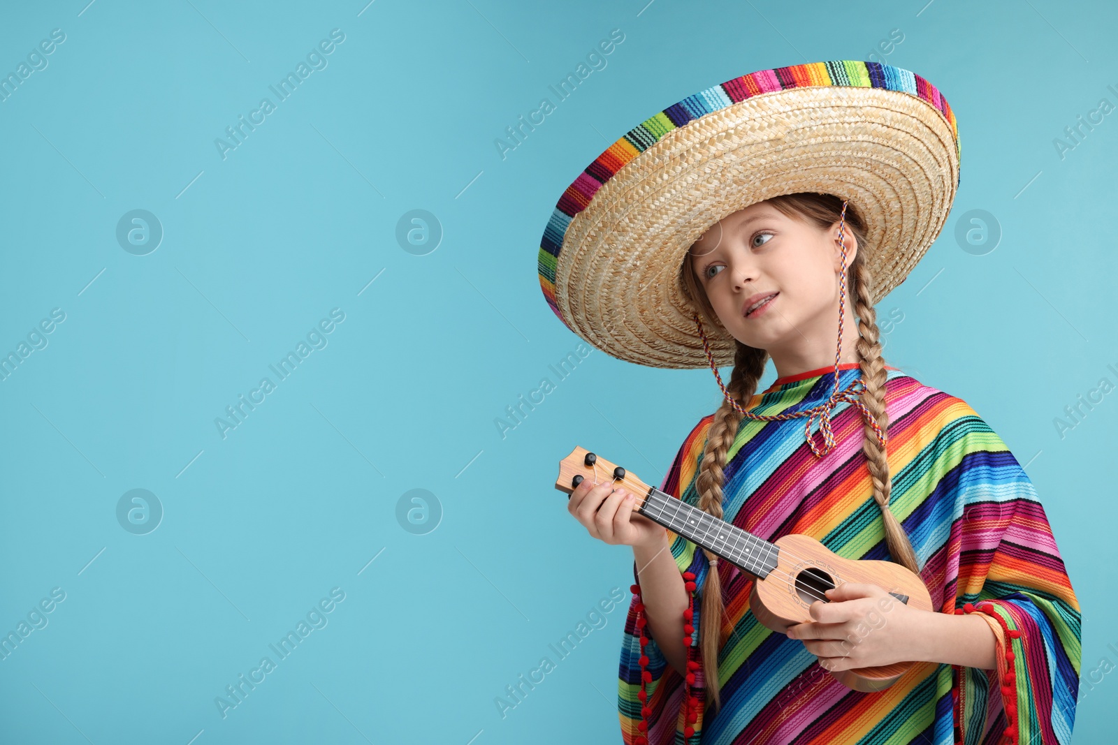 Photo of Cute girl in Mexican sombrero hat and poncho playing ukulele on light blue background. Space for text