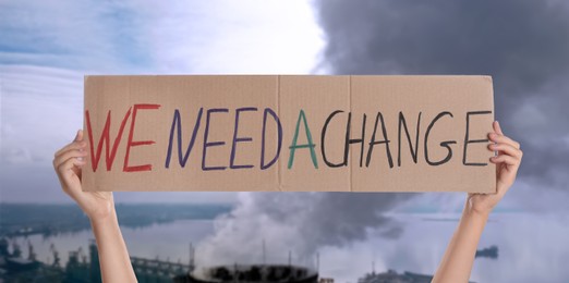 Protestor holding placard with text We Need A Change and blurred view of industrial factory on background. Climate strike