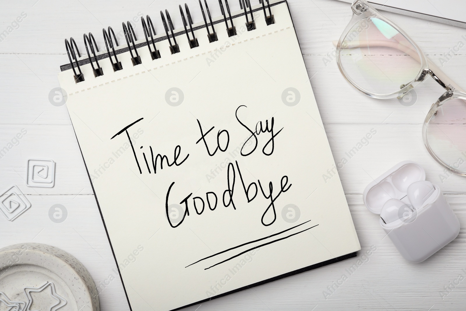 Image of Notebook with phrase Time to say Goodbye, eyeglasses and earphones on white wooden table, flat lay