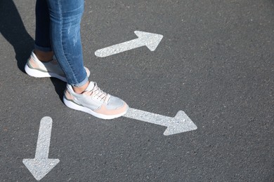Image of Choice of way. Woman walking to drawn mark on road, closeup. White arrows pointing in different directions