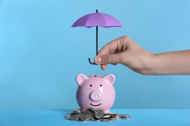 Photo of Woman holding small umbrella over piggy bank with coins on light blue background, closeup
