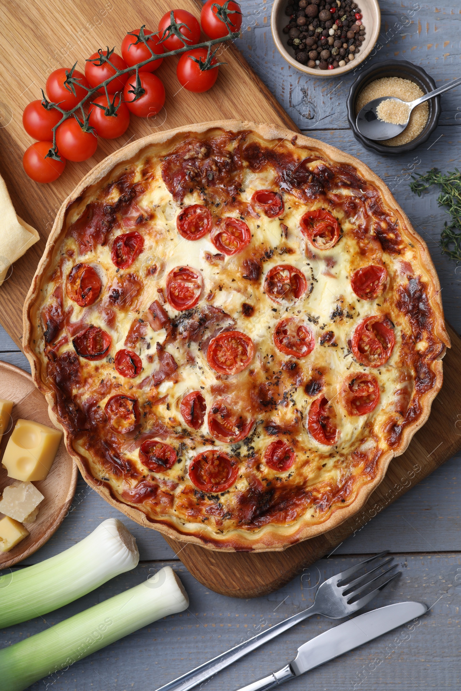 Photo of Delicious homemade prosciutto quiche, ingredients and cutlery on gray wooden table, flat lay