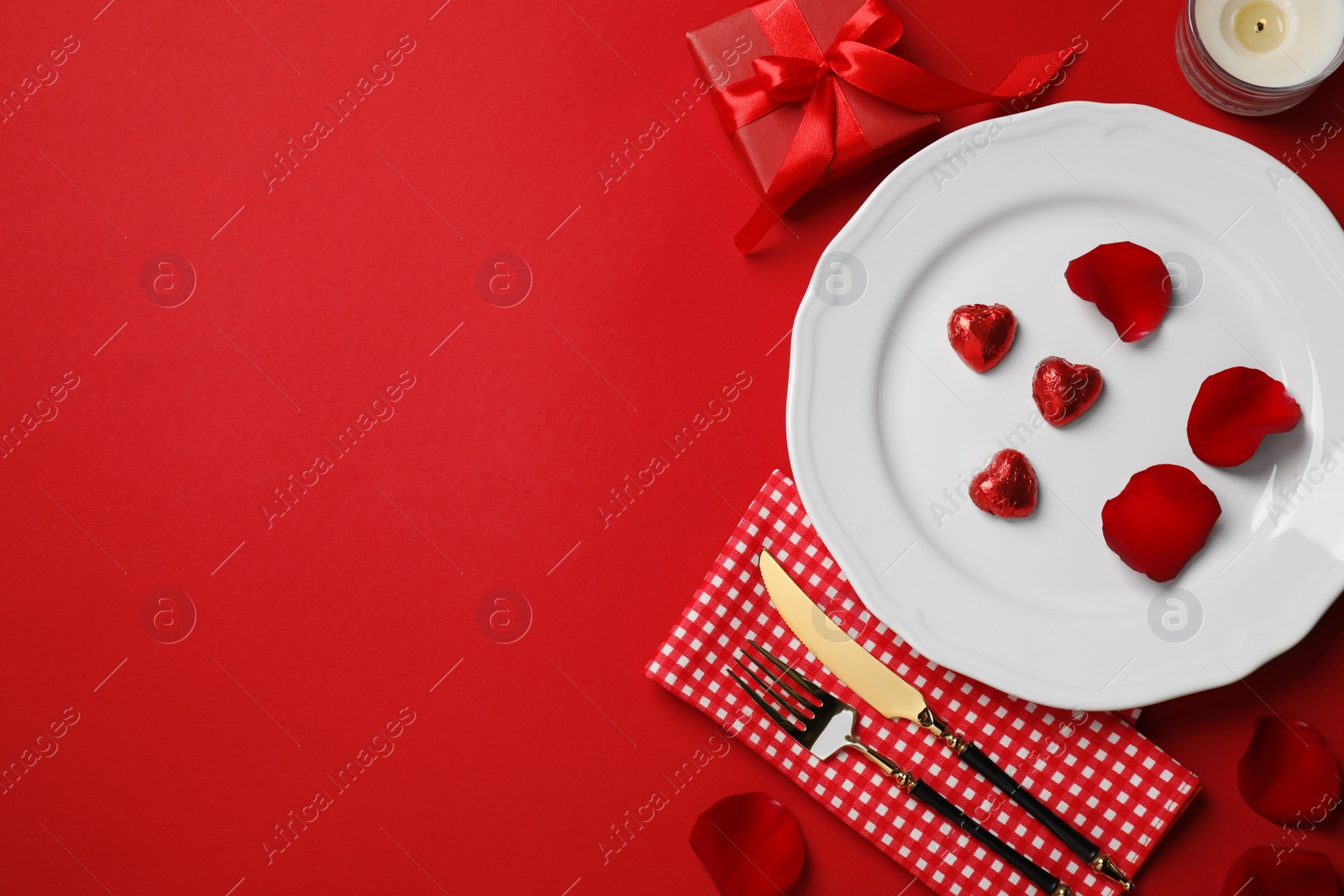 Photo of Beautiful table setting with gift box and rose petals on red table for romantic dinner, flat lay. Space for text
