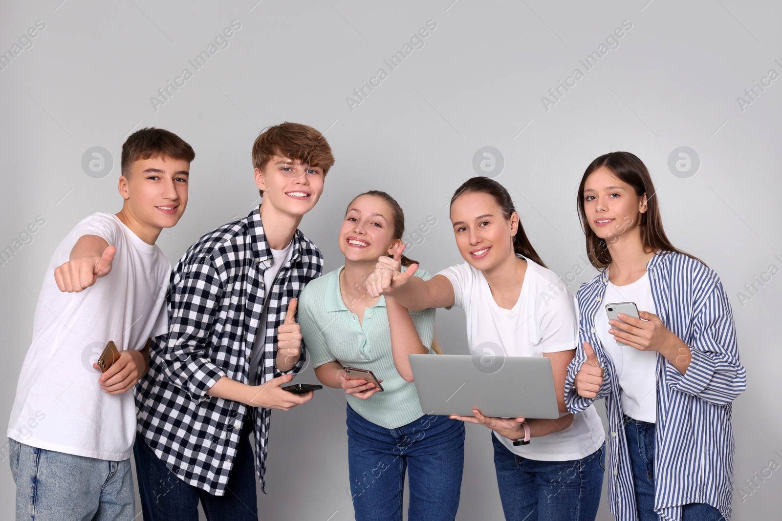 Photo of Group of happy teenagers with smartphones and laptop showing thumbs up on light grey background