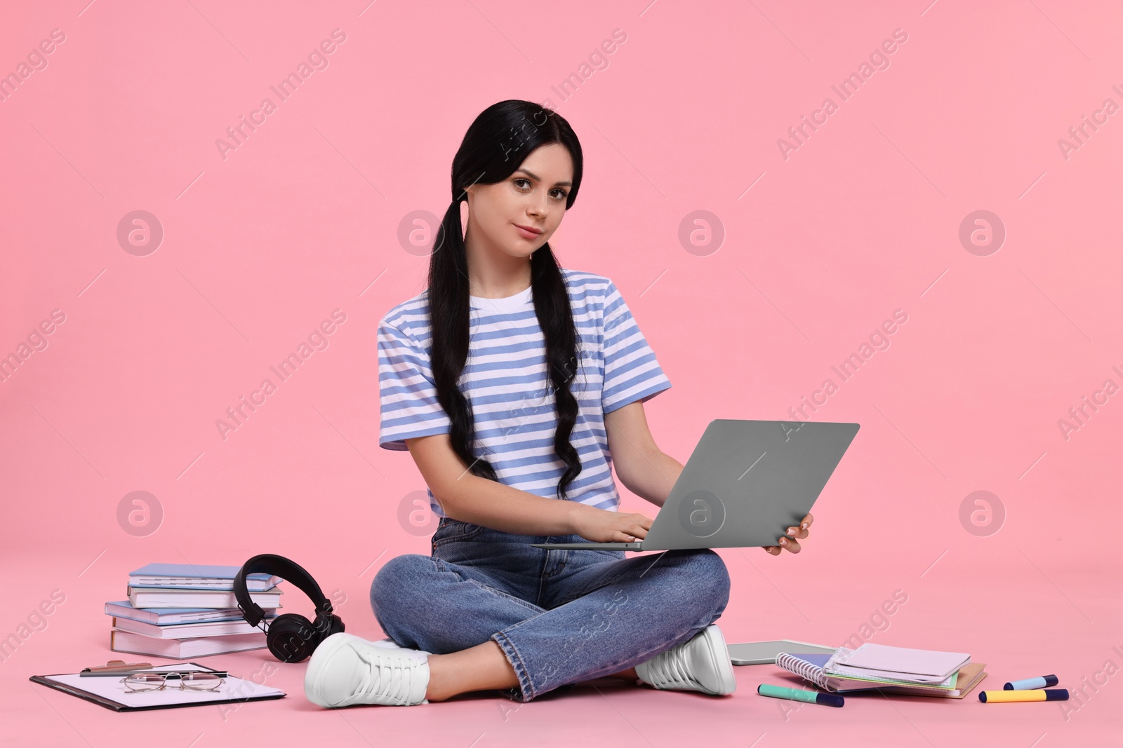 Photo of Student with laptop sitting among books and stationery on pink background