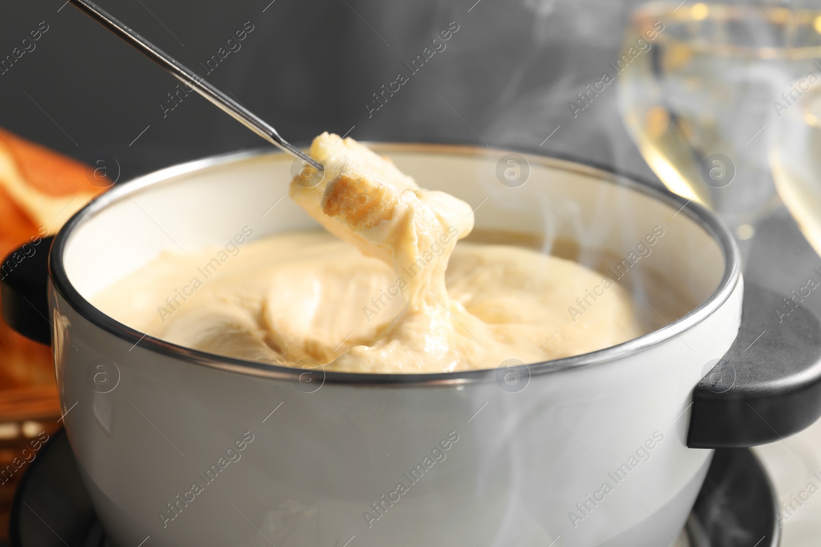 Photo of Dipping piece of bread into fondue pot with melted cheese at table, closeup