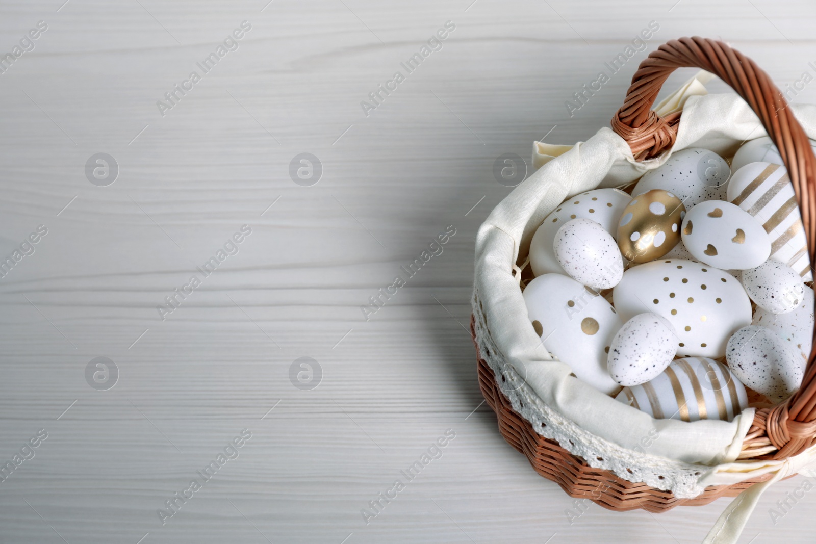 Photo of Wicker basket with festively decorated Easter eggs on white wooden table, above view. Space for text