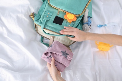 Photo of Woman packing baby accessories into maternity backpack on bed, top view
