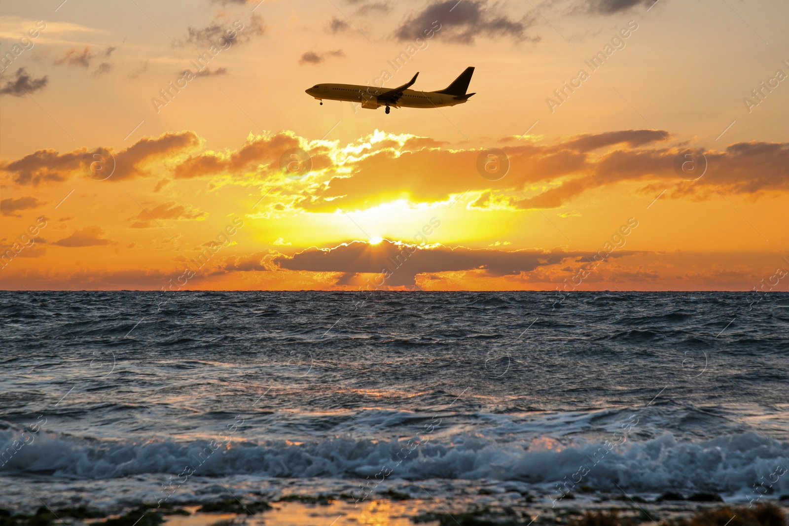 Image of Plane flying over sea during sunset. Sun shining through clouds