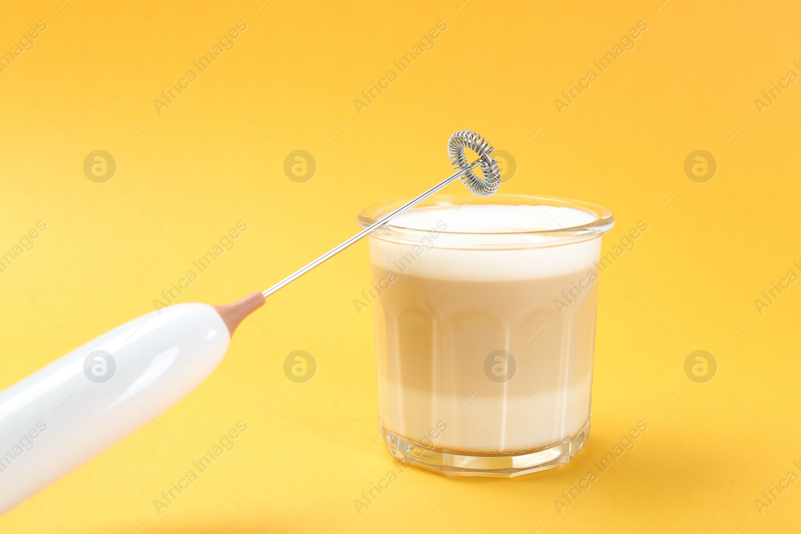 Photo of Mini mixer (milk frother) and tasty cappuccino in glass on yellow background