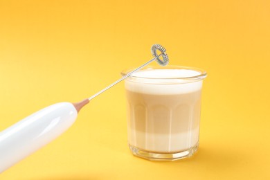 Mini mixer (milk frother) and tasty cappuccino in glass on yellow background