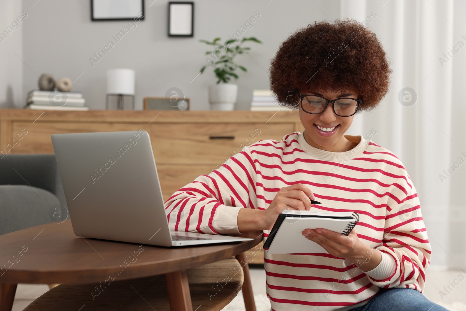 Photo of Beautiful young woman using laptop and writing in notebook at wooden coffee table in room