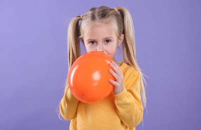 Photo of Cute little girl inflating orange balloon on violet background