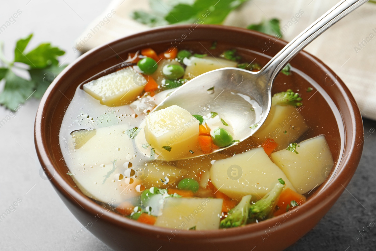 Photo of Spoon of fresh homemade vegetable soup over full bowl on grey table, closeup