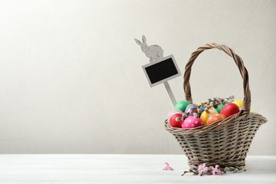 Photo of Colorful Easter eggs, flowers and tag in wicker basket on white wooden table against grey background. Space for text