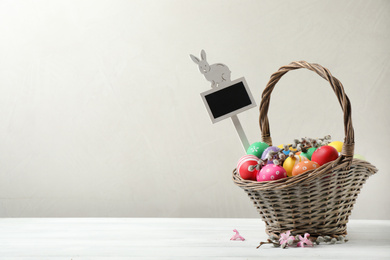 Colorful Easter eggs, flowers and tag in wicker basket on white wooden table against grey background. Space for text