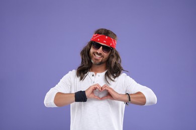 Photo of Stylish hippie man in sunglasses making heart with hands on violet background