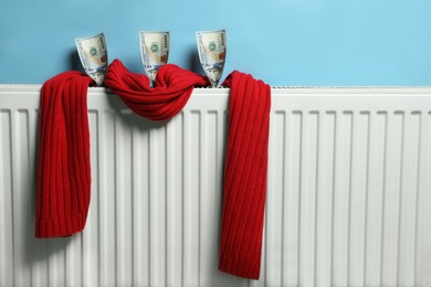 Modern radiator with knitted scarf and money near light blue wall indoors. Energy crisis concept
