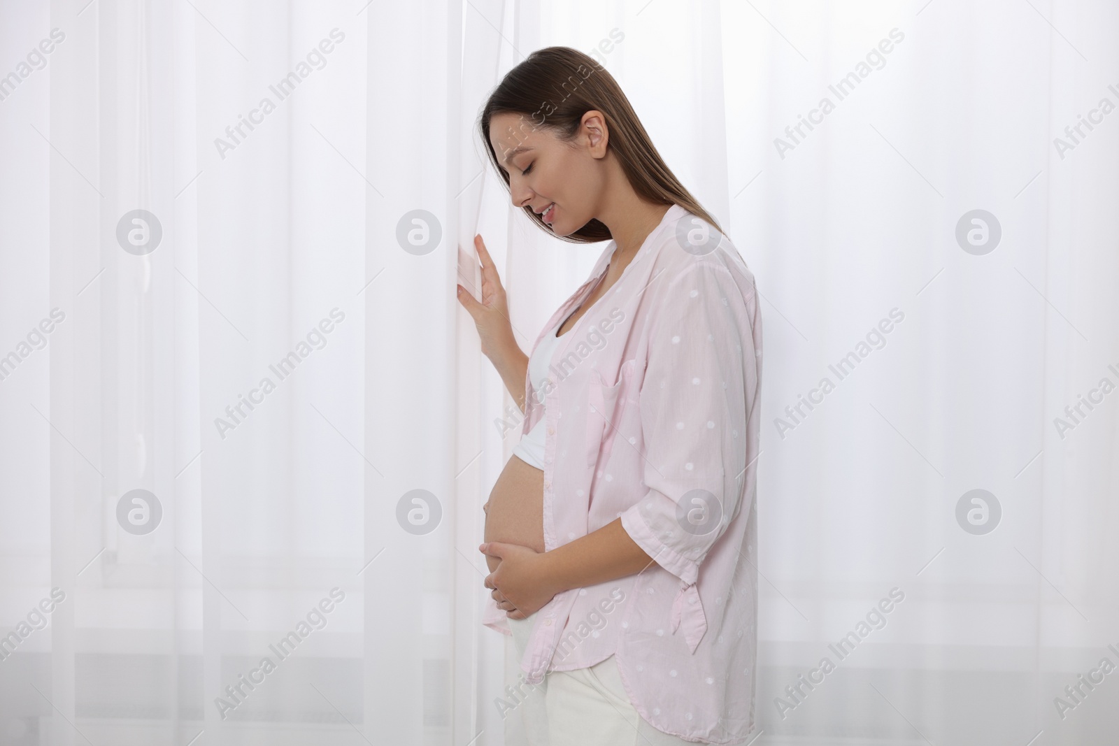 Photo of Beautiful pregnant woman in pink shirt near window indoors