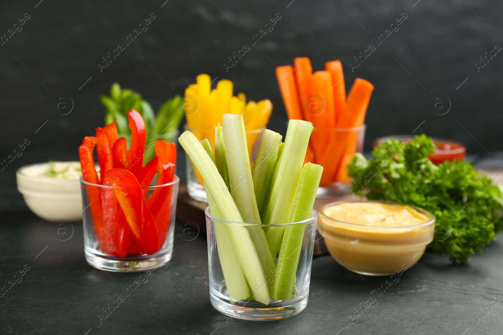 Photo of Celery and other vegetable sticks in glass bowls with dip sauce on black table