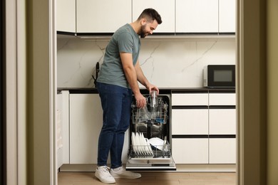 Smiling man loading dishwasher with glasses in kitchen