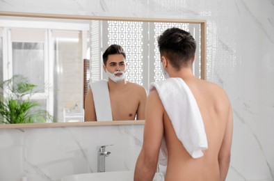 Handsome young man with shaving foam near mirror in bathroom