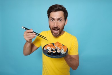 Photo of Emotional man holding plate with tasty sushi rolls and chopsticks on light blue background