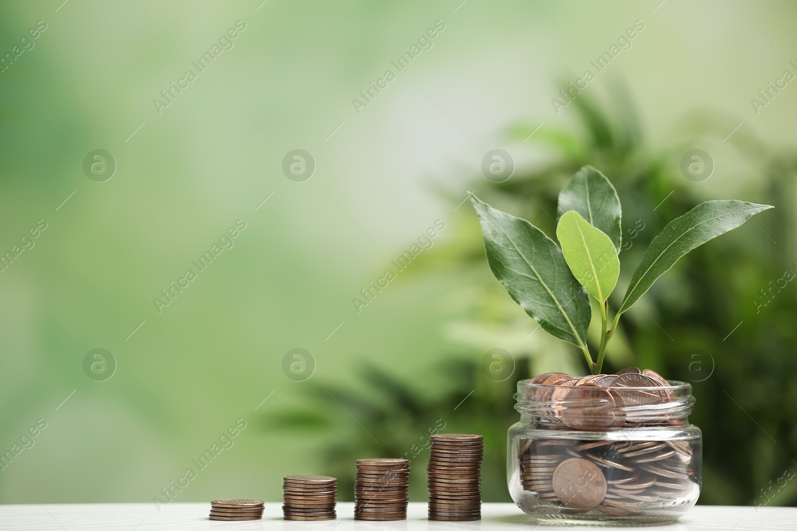 Photo of Glass jar and stacked coins with green plant on white table against blurred background. Space for text