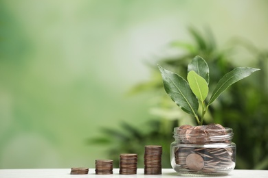 Glass jar and stacked coins with green plant on white table against blurred background. Space for text