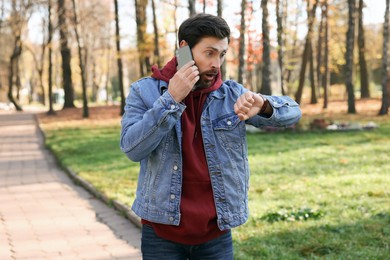 Photo of Emotional man talking by smartphone and checking time on watch in park. Being late concept