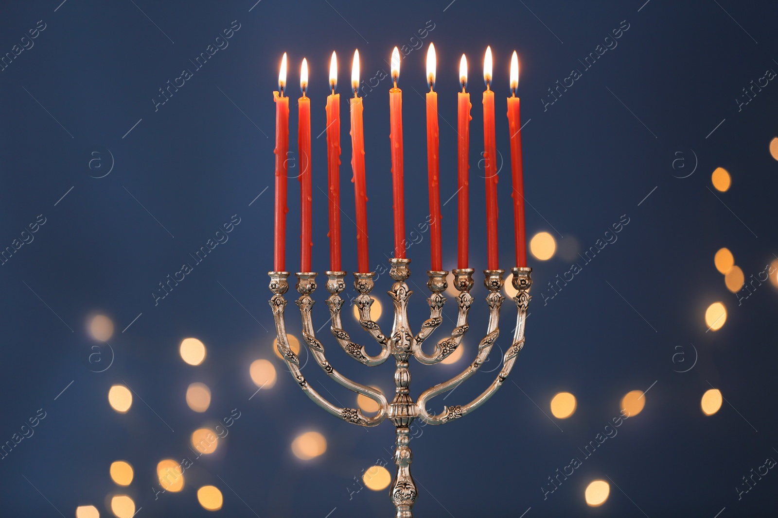 Photo of Silver menorah with burning candles against blue background and blurred festive lights. Hanukkah celebration