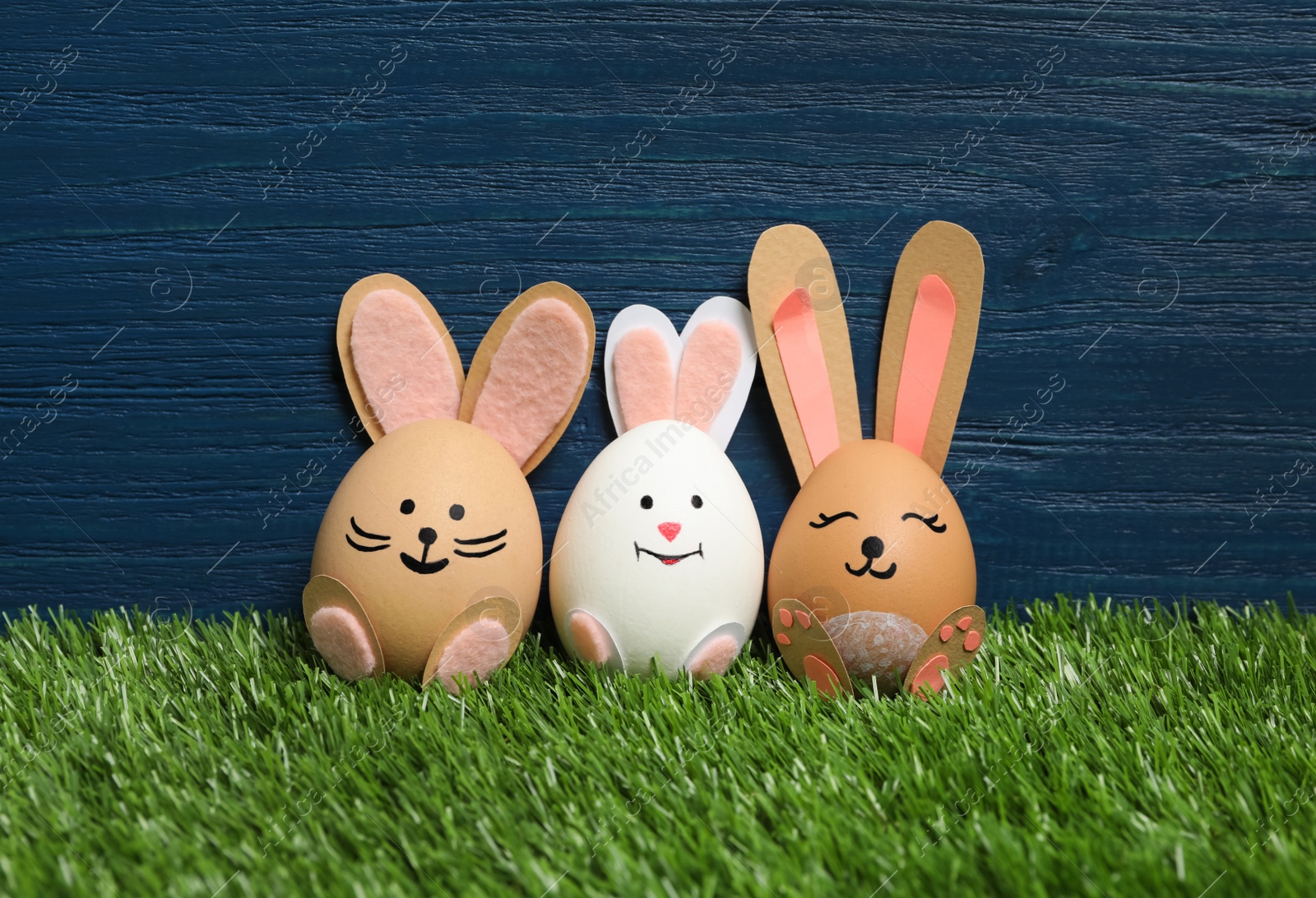 Photo of Bright Easter eggs as cute bunnies on green grass against blue wooden background