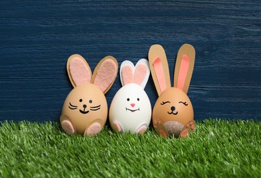 Photo of Bright Easter eggs as cute bunnies on green grass against blue wooden background