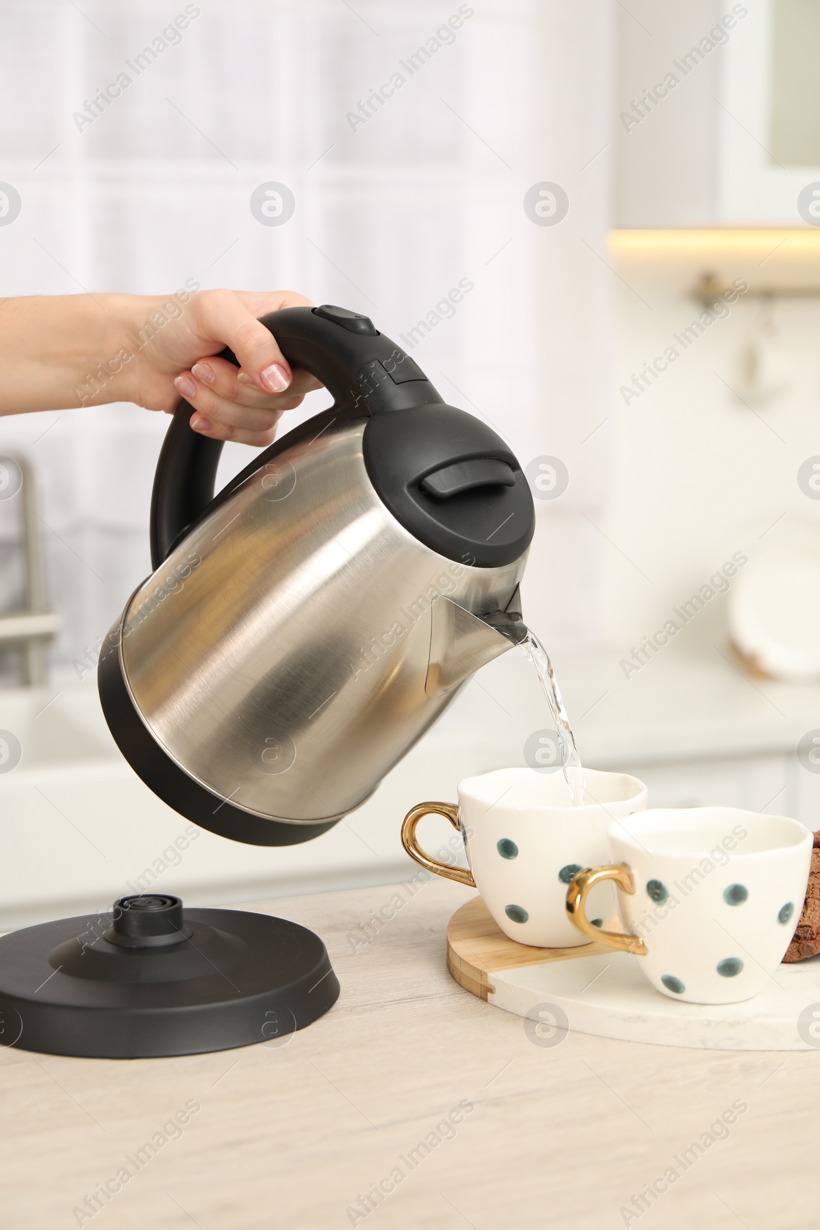 Photo of Woman pouring hot water from electric kettle into cup in kitchen, closeup