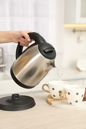 Photo of Woman pouring hot water from electric kettle into cup in kitchen, closeup