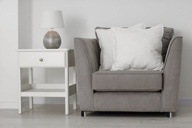 Photo of Soft white pillows on armchair, bedside table and lamp near light wall indoors