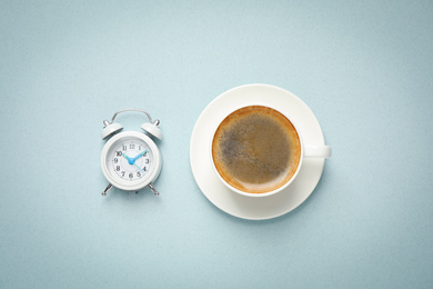 Cup of morning coffee and alarm clock on light background, flat lay