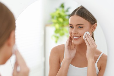 Photo of Beautiful woman removing makeup with cotton pad near mirror indoors