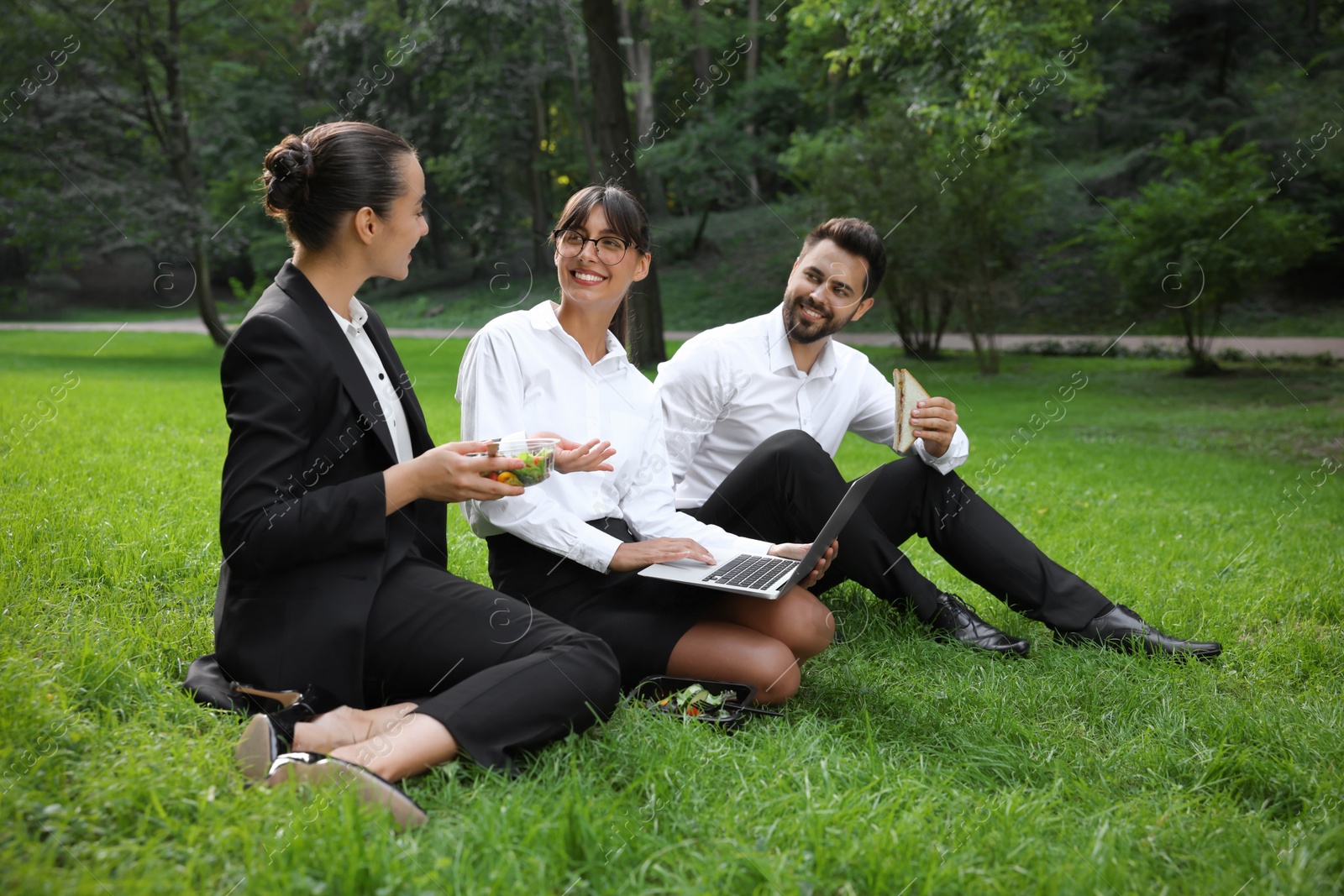 Photo of Happy colleagues with laptop having business lunch on green grass in park