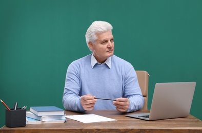 Photo of Portrait of senior teacher with laptop at table against green chalkboard