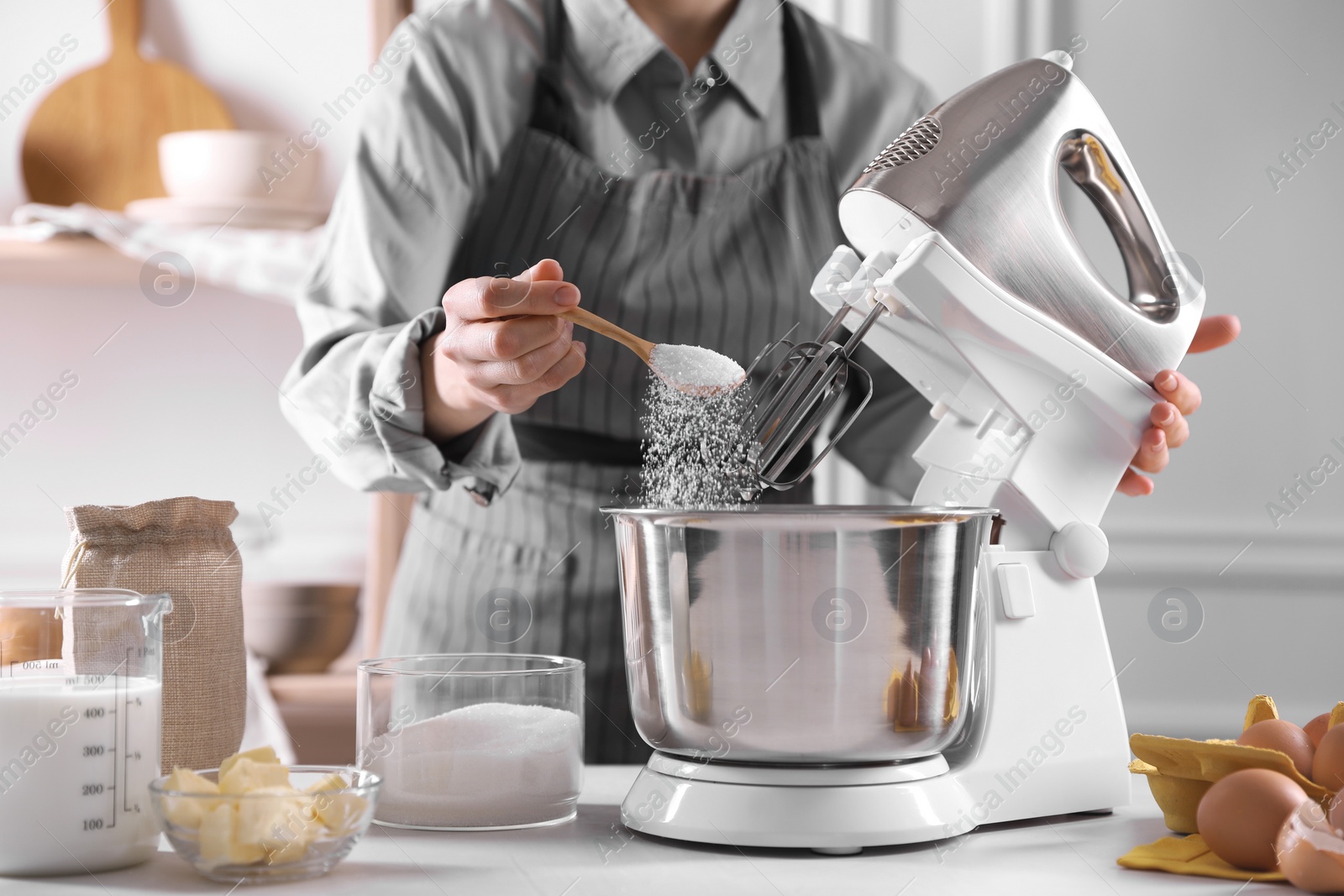 Photo of Woman adding sugar into bowl of stand mixer while making dough at table indoors, closeup