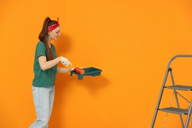 Photo of Happy designer with roller taking paint from container near orange wall